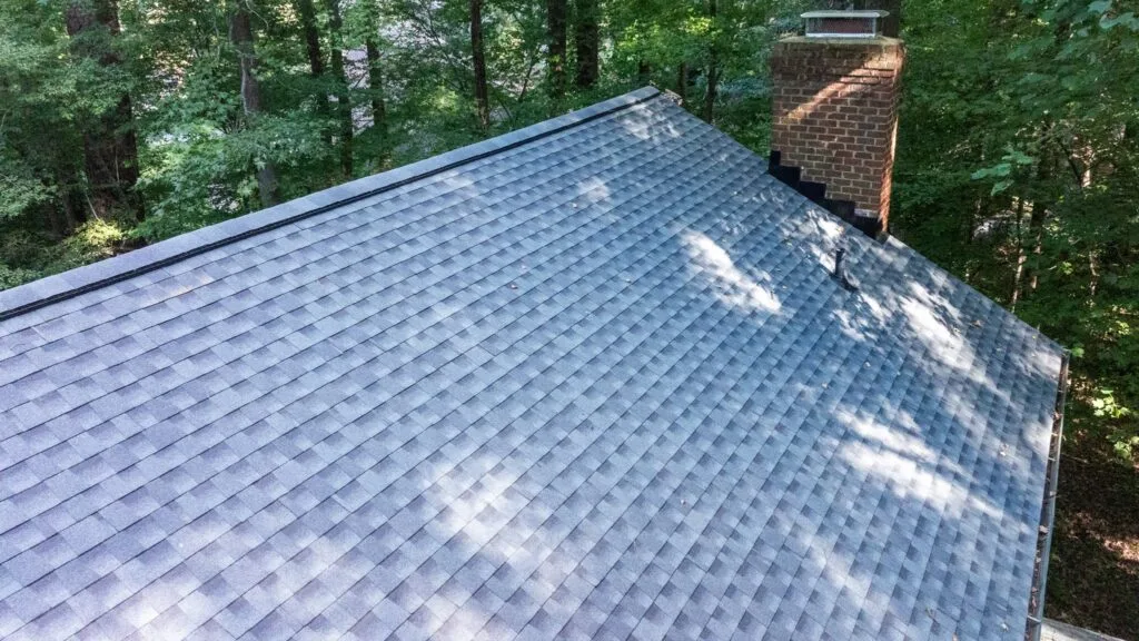 Aerial view of a grey asphalt shingle roof with a brick chimney surrounded by trees.