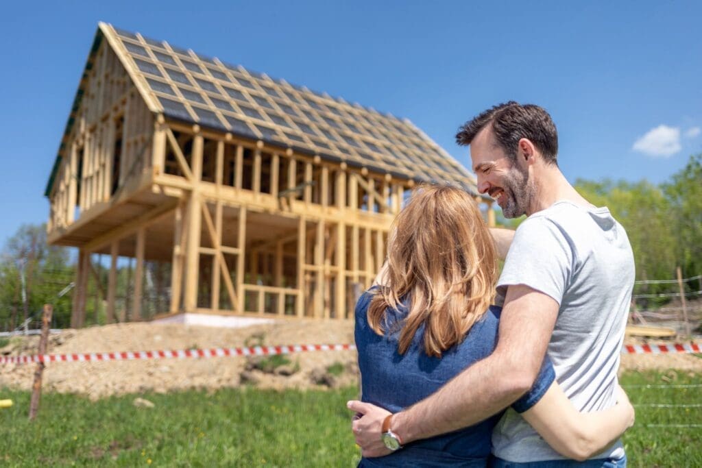 A smiling couple stands side-by-side with their arms around each other, looking at the wooden structural frame of a two-story house under construction in a field on a sunny day